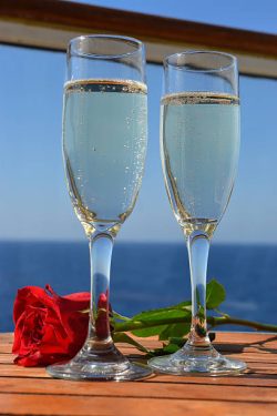 Two flutes of sparkling champagne and a red rose flower on a wooden table of a cruise ship with the blue ocean in the background.